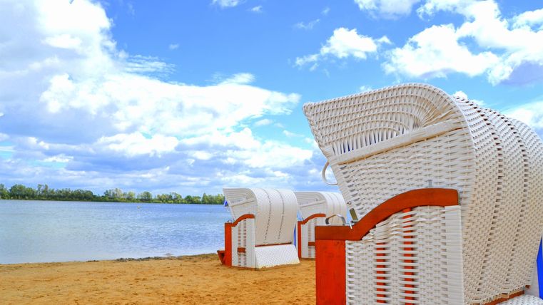 Strandstoelen staan aan de oever van een rustige waterloop. De lucht is blauw met enkele wolken.