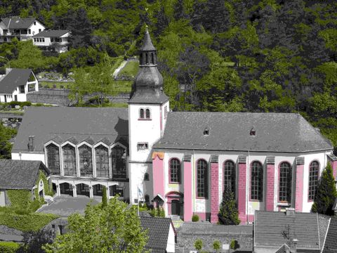 Eine schöne Kirche mit einem hohen Turm und bunten Fenstern. Sie ist von grünen Bäumen und Wohngebäuden umgeben.
