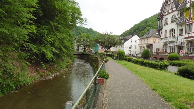 Eine malerische Straße entlang eines ruhigen Flusses, umgeben von Bäumen und Hügeln. Auf der anderen Seite stehen charmante Gebäude.