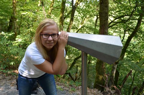 Eine junge Frau schaut durch ein Fernglas in einem Wald. Die Umgebung ist grün und lebendig.