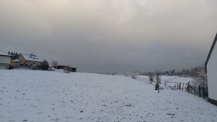 Een besneeuwd landschap met een licht bewolkte lucht. Op de achtergrond zijn enkele huizen en bomen te zien.
