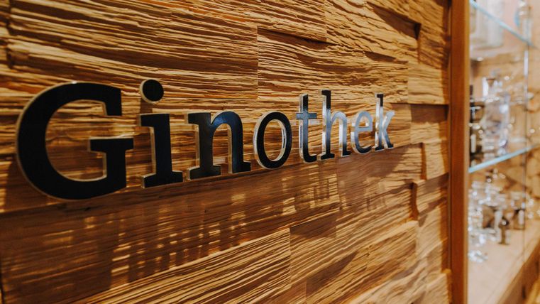 Eine Holzwand mit dem Schriftzug "Ginothek" in modernem Design. Im Hintergrund sind gläserne Regale mit verschiedenen Gin-Flaschen sichtbar.