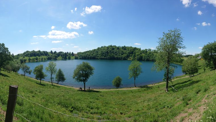A calm lake surrounded by green meadows and trees. The sky is clear and blue with a few white clouds.