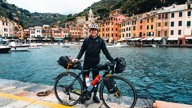 A cyclist stands at the edge of a picturesque harbor. Colorful buildings and calm water in the background create an inviting atmosphere.