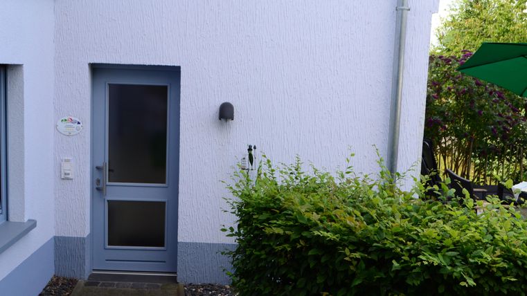 An entrance to a house with a gray door and a small front area. Next to the door is a green hedge and part of a garden is visible.