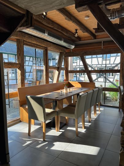 Een modern restaurant met een grote tafel en elegante stoelen. Grote ramen bieden veel natuurlijk licht en een uitzicht naar buiten.