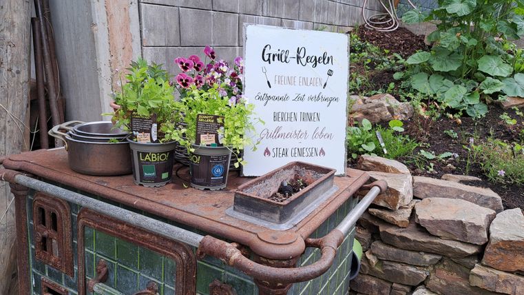 An old stove with flower boxes and a board that gives instructions for watering plants. In the background, green plants are visible.