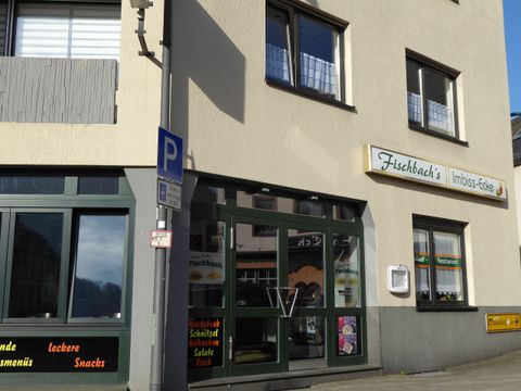 Ein Imbiss mit dem Namen "Fischbach's" in einem hellen Gebäude. Die Fenster sind groß und es gibt eine Markise sowie ein Parkschild vor der Tür.