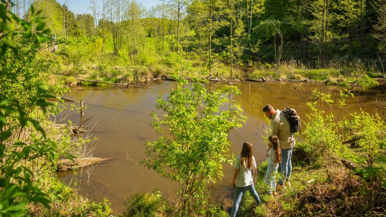 Een gezin staat aan een rustig water omgeven door weelderig groen. De zon schijnt en het is een mooie dag in de natuur.