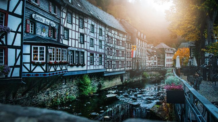 Half-timbered houses in Monschau by a river with autumn trees and sunshine.