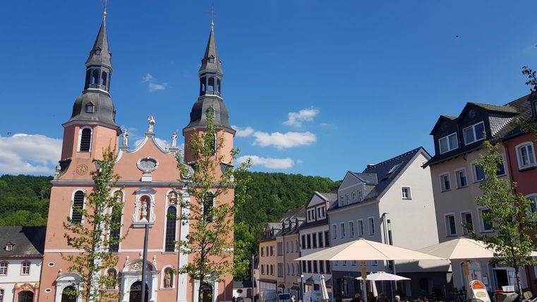 Ein schöner Platz mit einer markanten Kirche und modernen Gebäuden. Die Umgebung ist grün und einladend, ideal für einen entspannten Aufenthalt.