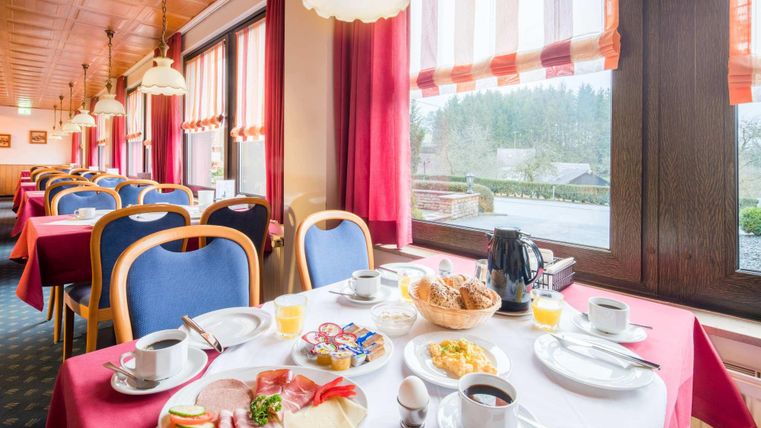 An inviting breakfast room with set tables and a diverse selection of dishes. In the background, there are windows with red curtains and a view of the landscape.