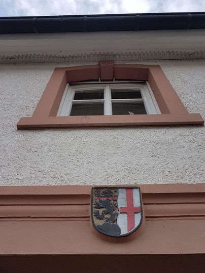 Ein Gebäude mit einem Fenster und einem Wappen darüber. Die Fassade ist hell und schlicht gestaltet.