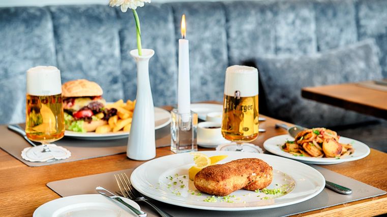 Een tafel in een restaurant met schnitzel, burger, frietjes en twee glazen bier. Een kaars en een bloem in een vaas staan op de tafel.
