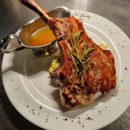 Een sappig stuk vlees met een bot, gegarneerd met verse kruiden. Daarbij is er een klein schaaltje met saus op een wit bord.