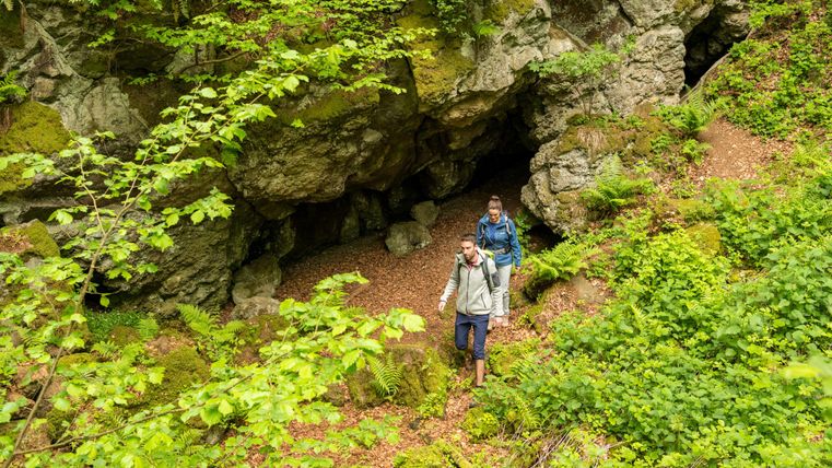 Zwei Wanderer gehen auf einem grünen Waldpfad an einer Felsenhöhle vorbei. Üppige Vegetation umgibt den Weg.