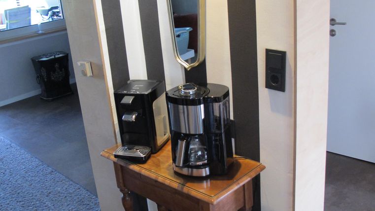 Ein moderner Kaffeekocher auf einer Holzkonsole steht an einer Wand mit gestreifter Tapete. Neben dem Kaffeekocher befindet sich ein Wandspiegel.