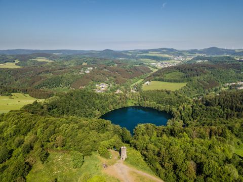 Eine schöne Landschaft mit einem klaren blauen See und grünen Wäldern. Im Hintergrund sind sanfte Hügel und kleine Ortschaften zu sehen.