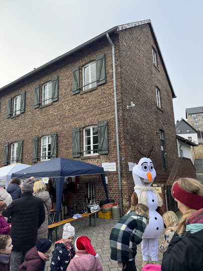 A festive event in front of an old building with a costume of a snowman. Many people, including children, are standing together and enjoying the atmosphere.