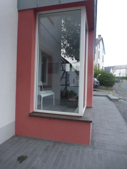 Eine Glasfront eines Gebäudes zeigt einen kleinen Raum mit einem weißen Stuhl. Die Umgebung ist urban und gut beleuchtet.