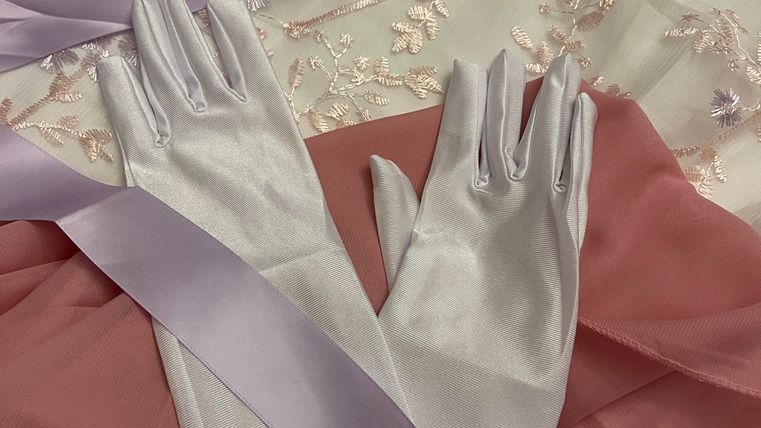 Elegant silver gloves lie on a delicate pink fabric. In the background, fine details of a light embroidered material can be seen.