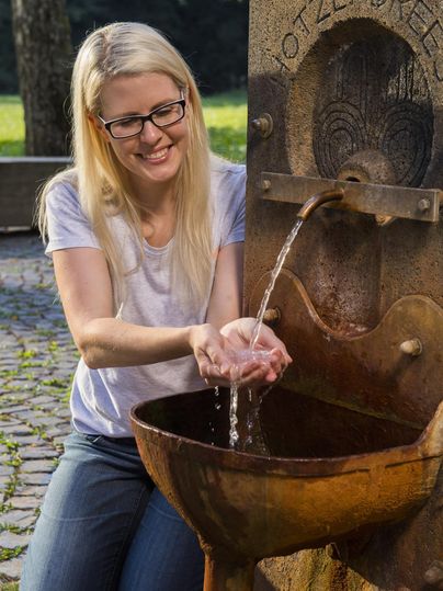 Eine Frau mit Brille steht an einem alten Brunnen und fängt mit ihren Händen sprudelndes Wasser auf. Im Hintergrund sind Bäume und ein ruhiger Weg zu sehen.