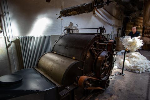 Eine alte Maschine in einer Fabrik, wahrscheinlich zur Verarbeitung von Baumwolle oder Wolle. Im Hintergrund steht ein Arbeiter, der mit einem Haufen Material beschäftigt ist.