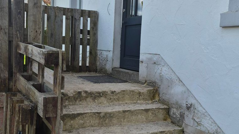 Eine Treppe aus Stein führt zu einer dunklen Tür in einem weißen Gebäude. Links ist ein Holzzaun zu sehen, und der Himmel ist mit einigen Wolken bedeckt.