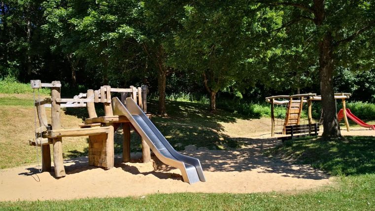 Een speelplaats met een glijbaan en een schommel. Het gebied is omgeven door bomen en heeft een zanderige ondergrond.