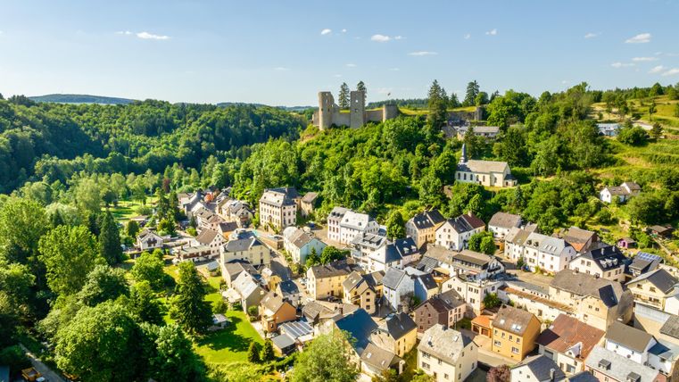 Eine malerische Dorfansicht mit kleinen Häusern und viel Grün. Im Hintergrund sind Burgruinen auf einem Hügel sichtbar.