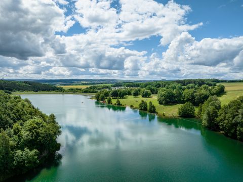 Ein ruhiger See umgeben von grünen Wäldern und Wiesen. Der Himmel ist klar mit großen, weißen Wolken.