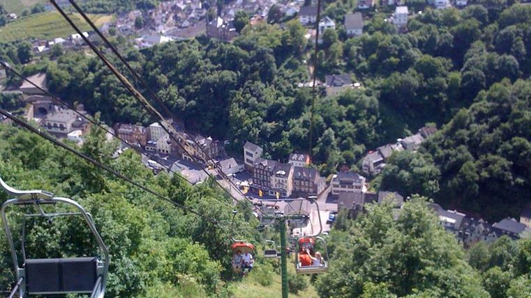Een kabelbaan rijdt over een groene Heuvel landschap met uitzicht op een klein dorp. De omgeving is omringd door bomen en biedt een schilderachtig uitzicht.