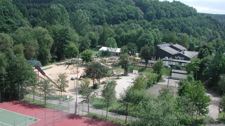 Aerial view of a holiday park with tennis court, playground, and wooded surroundings.
