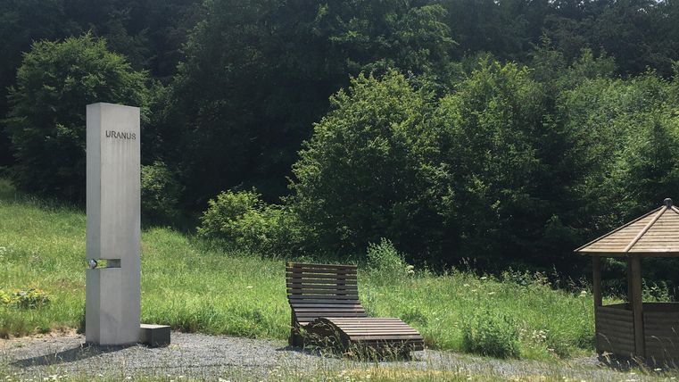 Eine sonnige Wiese mit einem modernen Duschgerät, einer Liege und einem Pavillon. Umgeben von grünen Bäumen bietet der Ort eine entspannende Atmosphäre.