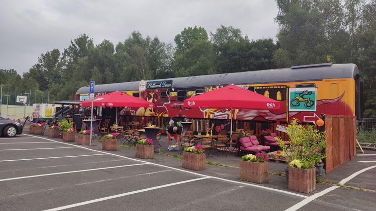 A colorful decorated restaurant cart with red umbrellas stands in a parking lot. Surrounded by plants and seating, it invites you to linger.