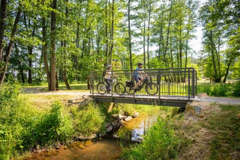 <p>Zwei Radfahrer überqueren einen kleinen Bach mittels einer Holzbrücke.Im Hintergund sieht man viele Laubbäume, die in Gruppen zusammen stehen. Die Sonne scheint</p>