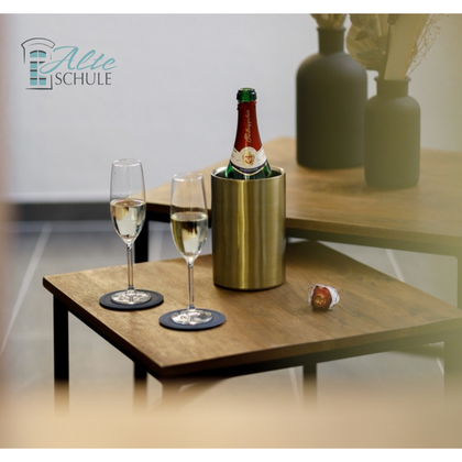 Zwei Gläser Sekt neben einem Flasche in einem Kühlbehälter auf einem Holztisch. Im Hintergrund sind dekorative Vasen zu sehen.