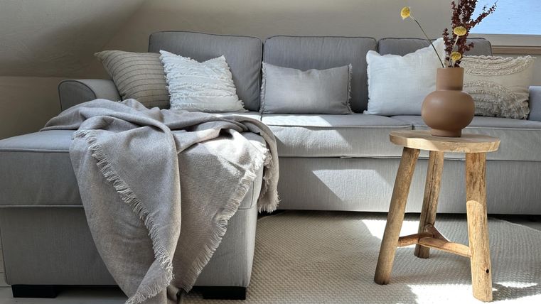 Een gezellige woonkamer met een grijze bank en meerdere kussens. Naast de bank staat een houten tafel met een vaas en verse bloemen.