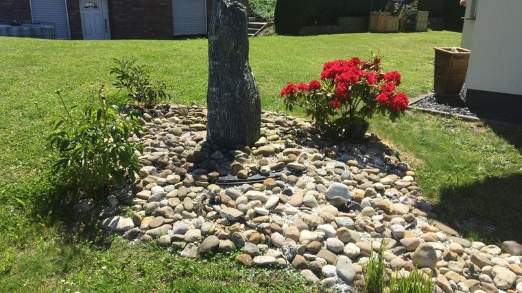 Een verzorgde tuin met een steen en kleurrijke bloemen. Rondom de steen liggen veel stenen op de grond.