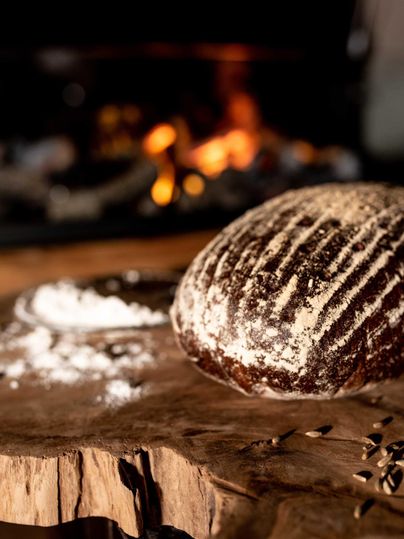 A freshly baked bread is lying on a wooden board. In the background, a fireplace can be seen.