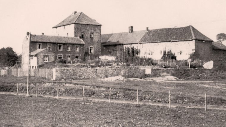 Een historische boerderij met een grote, aangelegde tuin. Op de achtergrond zijn nog meer gebouwen te zien, omgeven door een landelijke omgeving.