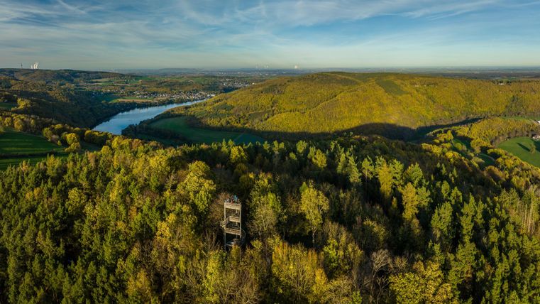 An impressive view of green hills and a river. In the midst of the trees stands an observation tower.