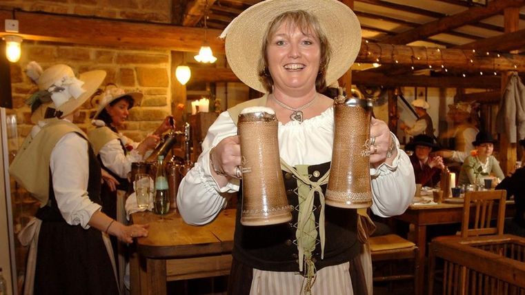 Eine traditionell gekleidete Frau lacht in die Kamera und hält zwei große Humpen Bier in den Händen. Im Hintergrund viele Gäste und weitere Kellnerinnen im Gasthaus der Birgeler Mühle.