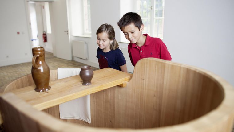 Zwei Kinder betrachten neugierig ein hölzernes Möbelstück. Auf dem Tisch stehen eine große und eine kleine Tontöpfe.