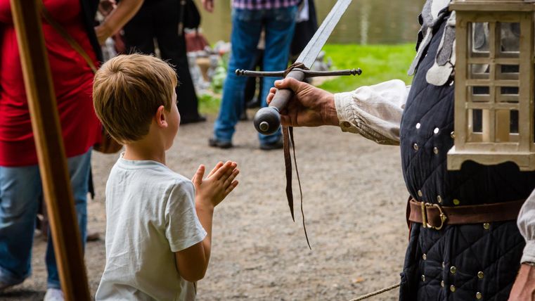 Een jongen staat met gevouwen handen en kijkt gespannen naar een zwaard dat een verklede man hem laat zien. Op de achtergrond zijn er meer personen en de natuur te zien.