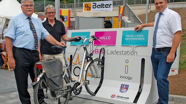Drei Männer stehen neben einer E-Bike-Ladestation. Ein Fahrrad ist an der Station angeschlossen. Im Hintergrund sind ein Zelt und ein Gebäude zu sehen.