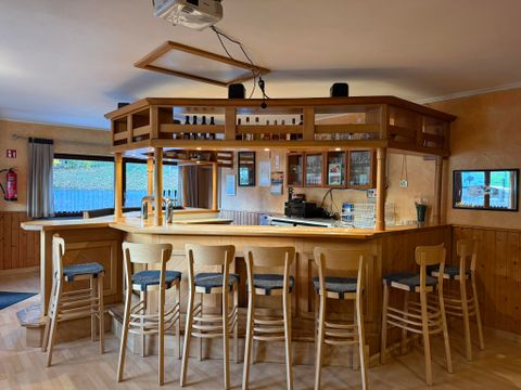 Eine gemütliche Bar mit Holzverkleidung und hohen Stühlen. Hinter der Bar sind Regale mit Getränken und ein Kaffeeautomaten sichtbar.