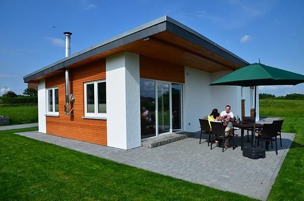A modern house with wooden cladding in a green environment. Outside, a group of people is sitting at a table under a sunshade.