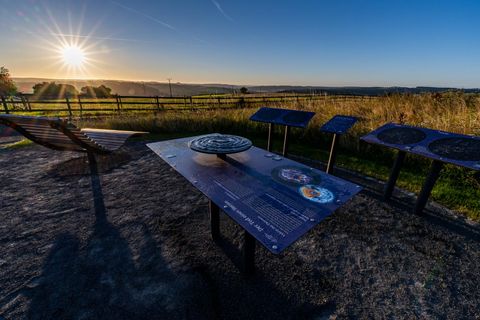 Ein Aussichtspunkt in der Natur mit einem weiten Blick auf die Landschaft. Die Sonne geht am Horizont auf, während informative Tafeln aufgestellt sind.