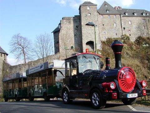 A small train stands in front of a castle. The surroundings are green with trees and historic stone buildings.